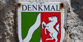 Blechschild mit Schriftzug "Denkmal" und Wappen Nordrhein-Westfalens, angeschraubt an einer Mauer