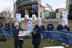 zwei Männer bei der Übergabe einer Box vor dem Landtag zusammen mit Unterstützer*innen mit Plakaten und Bannern